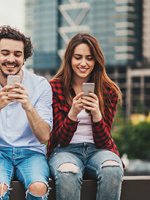 Zwei junge Menschen sitzen auf einer Mauer und schauen glücklich auf Ihre Smartphones.